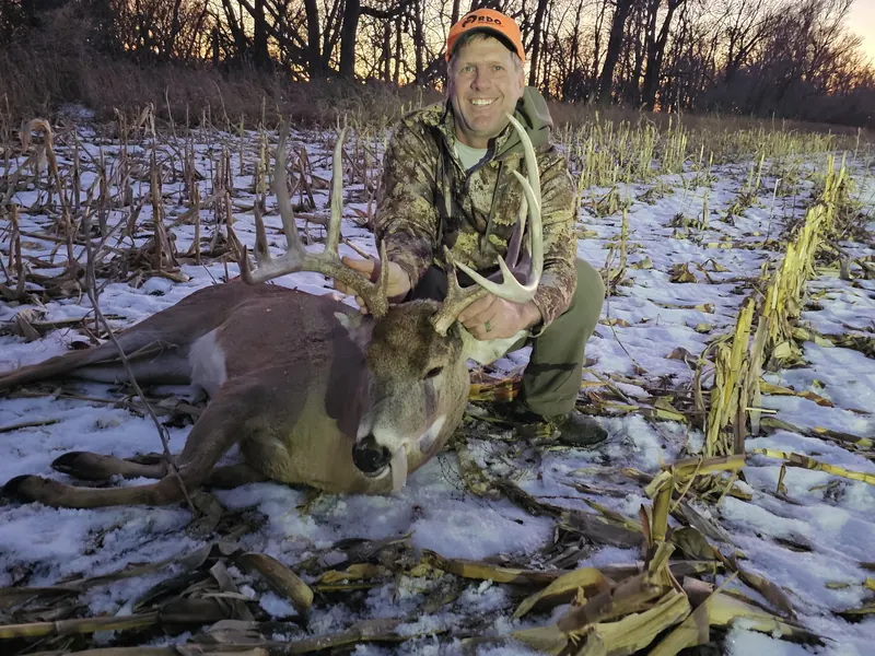 Hunter with a heavy whitetail buck in a snowy corn field at dusk