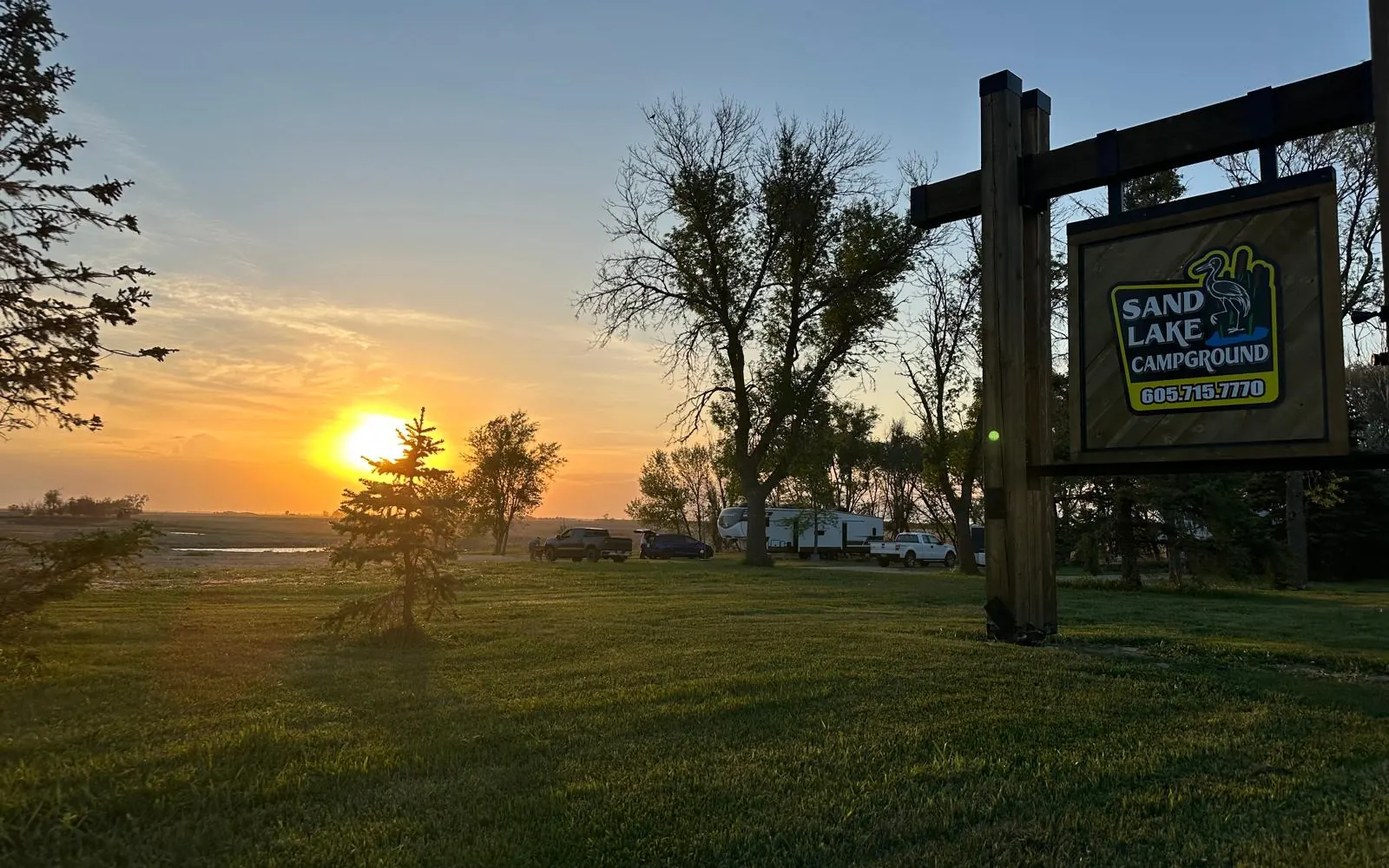 Sand Lake Campground sign at sunset with campers parked behind it